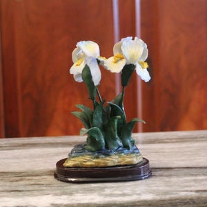Könnte beinhalten: Eine weiße Keramikfigur mit zwei Irisblüten mit grünen Blättern auf einem braunen Sockel. Die Blüten haben gelbe Zentren.