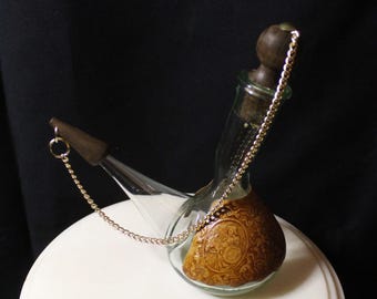 May include: A clear glass bottle with a brown, embossed leather base and a wooden stopper. A silver chain connects the stopper to the bottle. The bottle is on a white surface against a black background. The bottle is likely for perfume or essential oils.