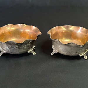 Op de afbeelding: Een paar kleine kommen met een gepolijste koperkleurige binnenkant en een doffe zilverkleurige buitenkant. Elke kom heeft een geschulpte, golvende rand en wordt ondersteund door drie sierlijke, getextureerde metalen voetjes die de basis verhogen.