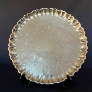 Op de afbeelding: Een rond, goudkleurig metalen dienblad met een geschulpte rand en een ingewikkeld gegraveerd bloemmotief over het oppervlak. Het dienblad staat rechtop op een kleine zwarte standaard, wat de reflecterende afwerking en het botanische ontwerp benadrukt.