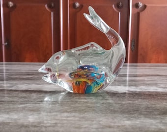 May include: A clear glass fish sculpture with a colorful interior. The fish has a curved tail and open mouth. The interior features swirls of blue, orange, and yellow. The sculpture is displayed on a gray surface.