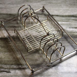 Op de afbeelding: Een rechthoekig zilverkleurig metalen afdruiprek met een doorzichtige acryl basis. Het rek bevat twee sets van drie verticale draadverdelers aan weerszijden en een centraal draadmandgedeelte voor het opbergen van borden of keukengerei.