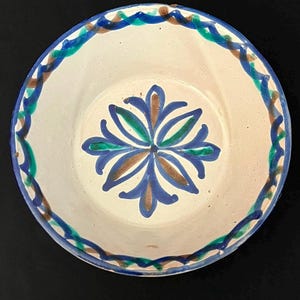 May include: A round ceramic bowl with an off-white base colour. The interior features a central floral motif painted in blue, green, and brown. The rim is decorated with a continuous wavy pattern in blue, green, and brown, encircling the bowl.