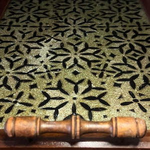 Op de afbeelding: Een rechthoekig houten dienblad met een donkerbruin frame en een handvat. De bodem van het blad toont een herhalend geometrisch patroon van goudkleurige, stervormige figuren tegen een contrasterende zwarte achtergrond.