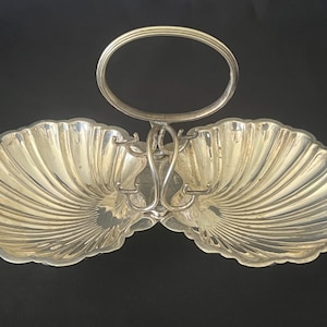 Peut inclure: Un plat de service en métal argenté composé de deux coupelles en forme de coquille avec des bords festonnés. Une poignée verticale centrale surmontée d'une boucle ovale relie les deux coupelles, offrant une structure pour transporter le plateau.