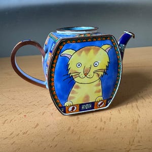 May include: A small, square-shaped metal teapot featuring a hand-painted yellow cat on a blue background. The teapot has a brown handle, a short spout, and a decorative border with orange, green, and blue geometric patterns around the cat illustration.