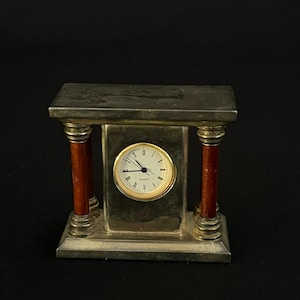 May include: A rectangular mantel clock featuring a silver-toned frame with two reddish-brown cylindrical columns on either side. The clock face is circular with a gold-toned bezel, white background, black Roman numerals, and the word Quartz printed.