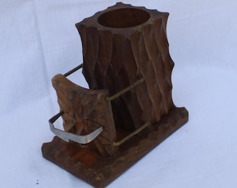 May include: A handcrafted wooden pen holder with a rustic, tree-trunk design. The brown holder features a carved texture and a metal clip for holding pens or pencils. The base is rectangular, and the top has a circular opening.