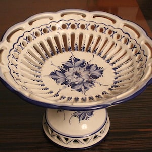 May include: A white ceramic fruit bowl with a blue floral pattern and a scalloped edge. The bowl is supported by a pedestal base, also decorated with blue accents. The bowl has a decorative, openwork design.