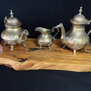 May include: A silver-toned metal tea set comprising a teapot, a lidded sugar bowl, and a creamer, each featuring a ribbed design and three ornate feet. A small clear glass thimble sits to the left of the set on a natural edge wooden board.
