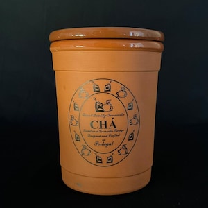 Op de afbeelding: Een cilindrische voorraadbus van terracotta met een bijpassend deksel. De voorzijde is voorzien van een cirkelvormige zwarte afbeelding met theezakjes-iconen en de tekst: Finest Quality Terracotta CHÁ Traditional Terracotta Storage Designed and Crafted in Portugal.