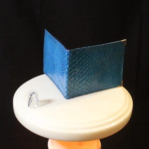 Puede incluir: Una cartera plegable con un patrón texturizado de piel de serpiente azul y detalles metálicos dorados en las esquinas. La cartera está colocada en posición vertical y abierta, mostrando una forma rectangular estructurada con una textura de escamas.