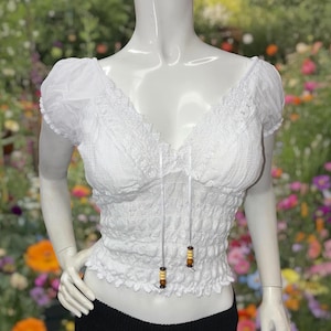 Puede incluir: Un top corto blanco de manga corta con un cuerpo fruncido texturizado, escote en V con ribete de encaje y dos finos lazos blancos que cuelgan del centro, cada uno terminado con pequeñas cuentas de madera marrón y amarilla.