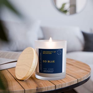May include: A lit, frosted glass candle with a wooden lid sits on a wooden table. The candle has a navy blue label with the text "GO BLUE" and "UNIVERSITY OF MICHIGAN". The candle is 11.5 oz and burns for 40 hours.