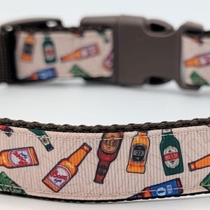 Peut inclure: Un collier pour chien avec une base en nylon marron et un revêtement en tissu beige clair. Le revêtement présente un motif répété de bouteilles de bière colorées. Le collier a une boucle en plastique marron et une boucle en métal argenté.