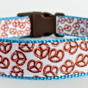Peut inclure: Un collier pour animal avec un ruban blanc orné d'un motif répétitif de bretzels marron, bordé de tissu bleu vif. Le collier comprend une boucle de fermeture en plastique marron et un anneau en métal argenté pour attacher une laisse.