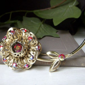 K&ouml;nnte beinhalten: Eine goldfarbene Brosche in Form einer Rose mit roten, schillernden Strasssteinen. Die Rose hat ein filigranes Design und einen gro&szlig;en Strassstein in der Mitte. Der Stiel der Rose ist ebenfalls mit Strasssteinen verziert.