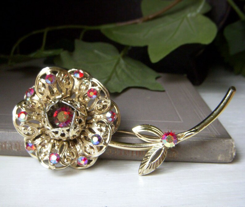 K&ouml;nnte beinhalten: Eine goldfarbene Brosche in Form einer Rose mit roten, schillernden Strasssteinen. Die Rose hat ein filigranes Design und einen gro&szlig;en Strassstein in der Mitte. Der Stiel der Rose ist ebenfalls mit Strasssteinen verziert.