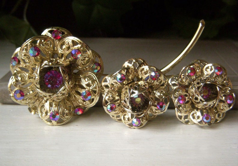 K&ouml;nnte beinhalten: Ein Set aus drei goldfarbenen Broschen in Blumenform mit schillernden rosa Strasssteinen.