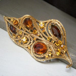 May include: A gold tone brooch with a leaf design. The brooch is decorated with amber and gold colored rhinestones.