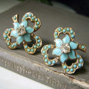 May include: A pair of gold-tone metal earrings shaped like four-leaf clovers. Each earring features light blue enamel petals, a central clear rhinestone, and a border of small, round, light blue beads set along the clover-shaped metal frame.