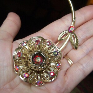 K&ouml;nnte beinhalten: Eine goldfarbene Brosche in Form einer Blume mit roten Strasssteinen. Die Blume hat ein zartes, filigranes Design und ist an einem Stiel mit Bl&auml;ttern befestigt.