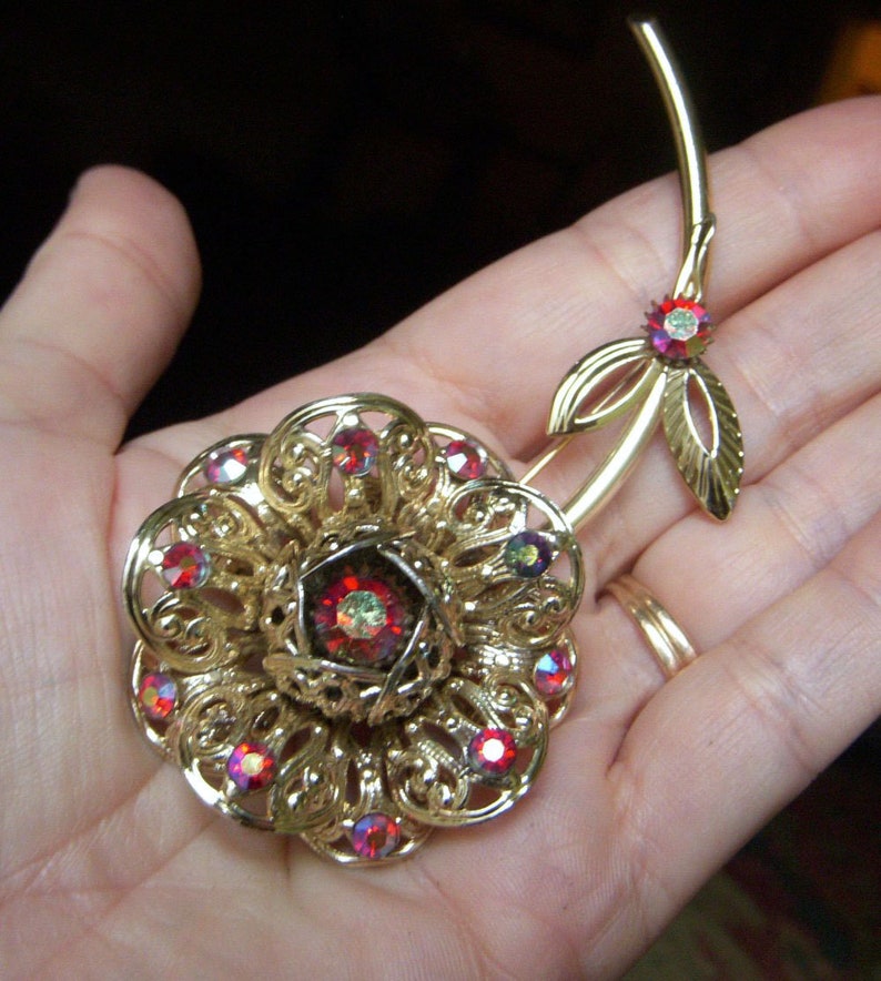 K&ouml;nnte beinhalten: Eine goldfarbene Brosche in Form einer Blume mit roten Strasssteinen. Die Blume hat ein zartes, filigranes Design und ist an einem Stiel mit Bl&auml;ttern befestigt.