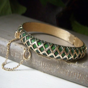 May include: A gold-tone hinged bangle bracelet featuring a raised lattice pattern filled with deep green enamel. The bracelet includes a thin gold-tone safety chain with a spring ring clasp attached to the side, showcasing a textured interior finish.