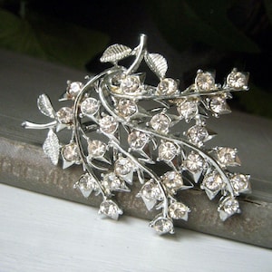 May include: Silver-toned brooch in a floral design, featuring delicate leaves and sparkling clear crystals. The brooch has a detailed, three-dimensional appearance, with the crystals adding a touch of elegance. The brooch is likely made of metal.