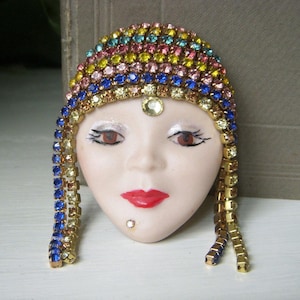 May include: A vintage face-shaped brooch with a colorful jeweled headdress. The face has red lips, brown eyes, and a small beauty mark. The headdress is adorned with a variety of colorful rhinestones, including pink, blue, yellow, and green.