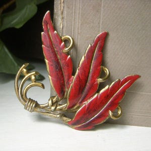 May include: Vintage brooch featuring three red enameled leaves with gold trim and accents. The leaves have a textured appearance with dark red veining. The brooch has a gold-toned metal base with decorative swirls and loops.