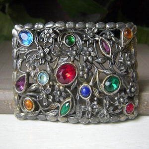May include: A silver-toned metal brooch with a floral design and colorful rhinestones. The rhinestones are red, blue, green, and purple.
