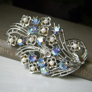 May include: Silver-toned brooch featuring a floral design with iridescent blue and clear crystals, and small faux pearls. The brooch has a detailed, openwork metal design, and is likely vintage or antique.