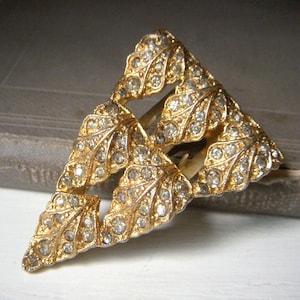 May include: A triangular brooch featuring a gold-tone metal frame shaped like a cluster of leaves. The surface of each leaf is textured and encrusted with numerous small, clear, sparkling rhinestones that catch the light across the entire piece.