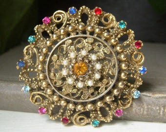 Könnte beinhalten: Vintage goldfarbene Brosche mit kreisförmigem Design. Die Brosche hat ein aufwendiges Filigranmuster, verziert mit farbigen Strasssteinen in Rot, Blau, Rosa und Grün. Die Mitte hat einen orangefarbenen Edelstein, umgeben von Kunstperlen.