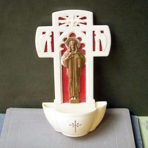 May include: A cream-colored wall-mounted holy water font featuring a central bronze-toned figure of Jesus with a halo, set against a red background. The cross-shaped frame includes angel silhouettes and a dove, with a small cross symbol on the basin.