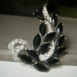 May include: A silver brooch with a design of black marquise-shaped stones set in a swirling pattern. The brooch is made of metal and rhinestones.