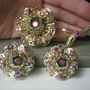 K&ouml;nnte beinhalten: Ein Set aus goldfarbenem Schmuck mit einer gro&szlig;en Blumenbrosche und passenden Ohrringen. Der Schmuck ist mit roten, schillernden Strasssteinen verziert.