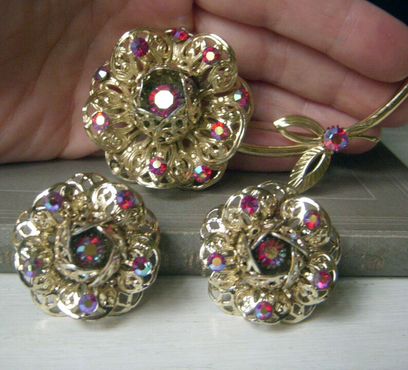 K&ouml;nnte beinhalten: Ein Set aus goldfarbenem Schmuck mit einer gro&szlig;en Blumenbrosche und passenden Ohrringen. Der Schmuck ist mit roten, schillernden Strasssteinen verziert.