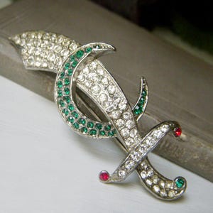 May include: A vintage brooch with a silver-tone metal base, adorned with clear and green rhinestones. The design features a stylized letter or symbol, with red and green accents. The brooch has a pin closure on the back.