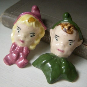 May include: Two vintage ceramic figurines, one with a pink hat and dress, the other with a green hat and collar. Both have painted faces with delicate features, including black eyes and pink lips. The figurines are likely decorative collectibles.