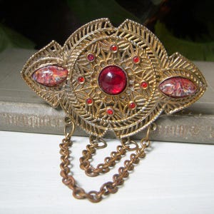May include: Vintage gold-tone brooch with a filigree design. The brooch features a large, round, red center stone and smaller red accents. Two oval-shaped stones with a marbled appearance are on either side. Two chains hang from the bottom.