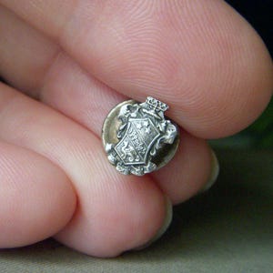 May include: A small, antique silver-toned button with a detailed crest design. The crest features a shield with stars, a lion, and ornate scrollwork. A small crown sits atop the crest. The button has a slightly tarnished appearance, suggesting age.