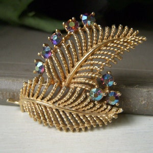 May include: A gold-tone brooch in the shape of a fern leaf with iridescent purple, blue, and green rhinestones.