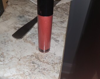 Pode incluir: Um tubo de gloss labial vermelho com uma tampa preta. O gloss tem um acabamento brilhante.