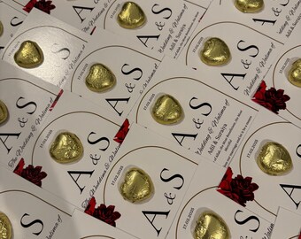 May include: Arrangement of wedding favor cards with gold foil-wrapped heart-shaped chocolates. Each card features the initials "A & S", the wedding date, and floral accents. The cards are white with black text.