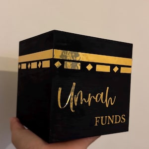 May include: Black wooden box with gold trim and the words "Umrah Funds" written on the front.