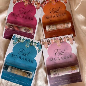 Könnte beinhalten: Vier Eid Mubarak-Grußkarten in Pink, Orange, Blau und Lila. Jede Karte hat ein Laternen-Design und eine aufgerollte Banknote. Die Karten zeigen den Text "Eid Mubarak" und eine Nachricht.