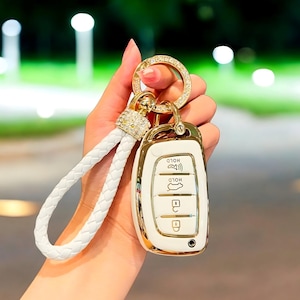 May include: A white and gold car key fob with a braided white leather strap and a gold ring with rhinestones. The key fob has gold trim and buttons for locking and unlocking the car.