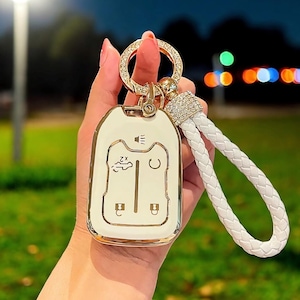 Può includere: Portachiavi per auto bianco con finiture dorate e un portachiavi intrecciato bianco. Il portachiavi ha una piccola icona dell'altoparlante, un'icona dell'auto e due pulsanti. Il portachiavi ha un anello dorato e un abbellimento di cristallo.